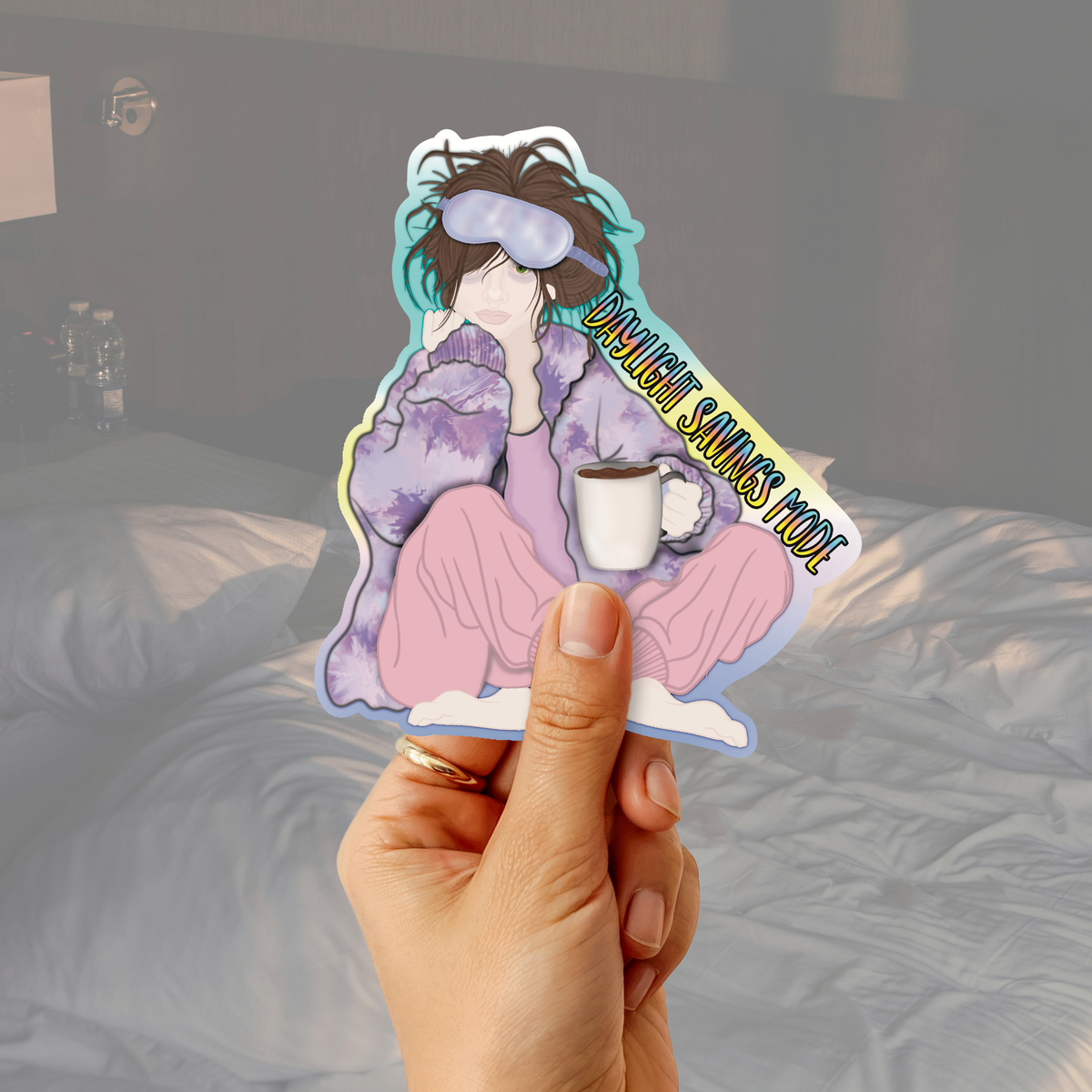 A hand holds a die-cut sticker featuring an illustrated person in a purple robe and pink pants holding a mug, with the text 'DAYLIGHT SAVINGS MODE'. The background is a softly blurred bedroom scene with a bed and bedside lamp.