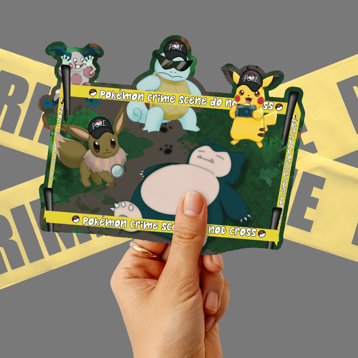 A hand holds a rectangular collectible card featuring Pokémon characters in a crime scene setup, with yellow police tape graphics and text reading 'Pokémon crime scene do not cross.' The background is a gray studio backdrop with blurred yellow tape graphics.