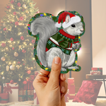 A hand holds a die-cut sticker or decal shaped like a gray squirrel wearing a red Santa hat and green holiday sweater, with a blurred Christmas tree and gift-wrapped boxes in the background.