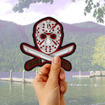 A hand holds a die-cut sticker featuring a hockey mask with crossed knives behind it, set against a blurred lake and forest background.