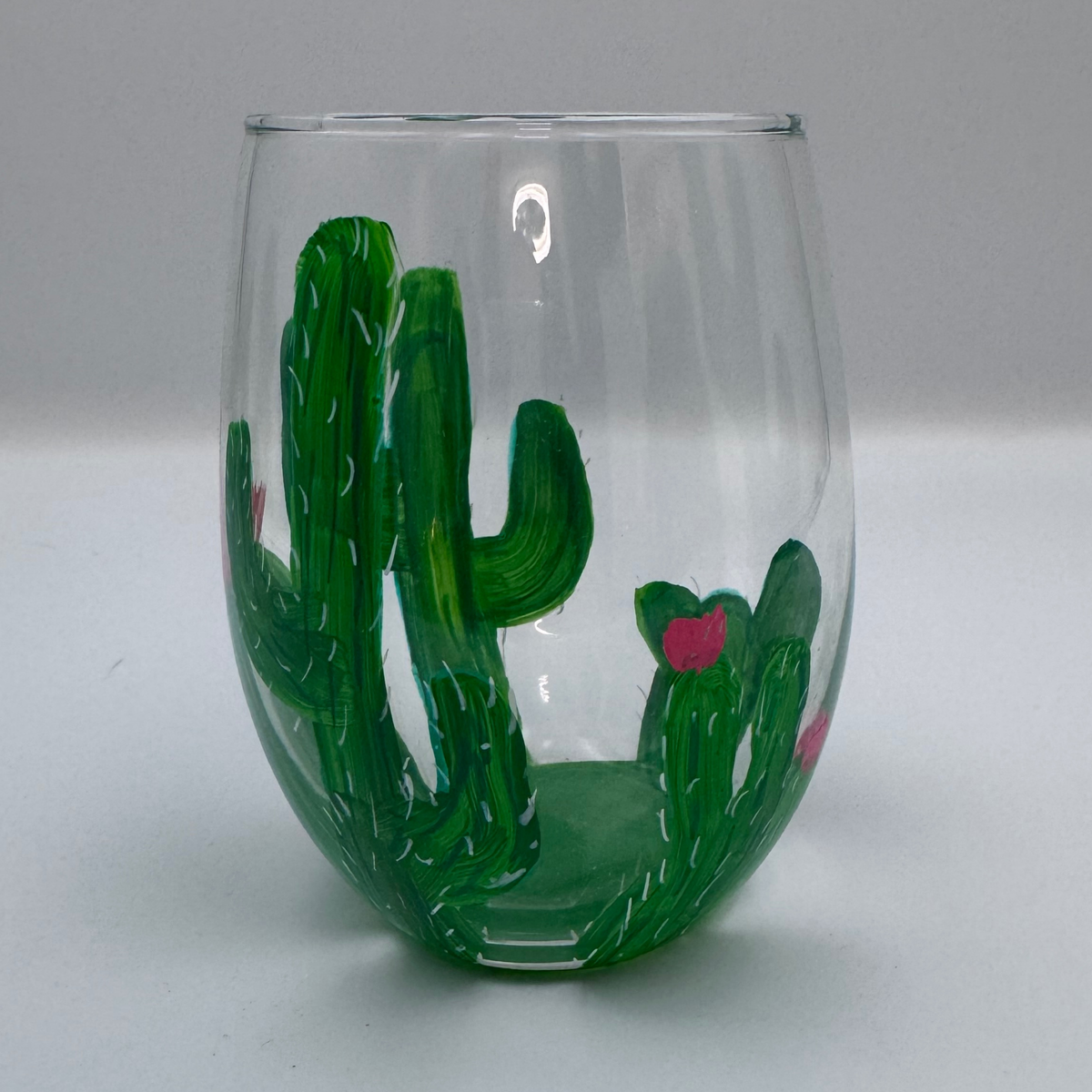 A clear stemless wine glass decorated with painted green cacti and small pink flowers, shown against a plain light gray background.