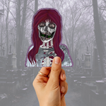 A hand holds a die-cut sticker featuring a skeleton character with long dark purple hair and a white shirt, set against a foggy cemetery background.