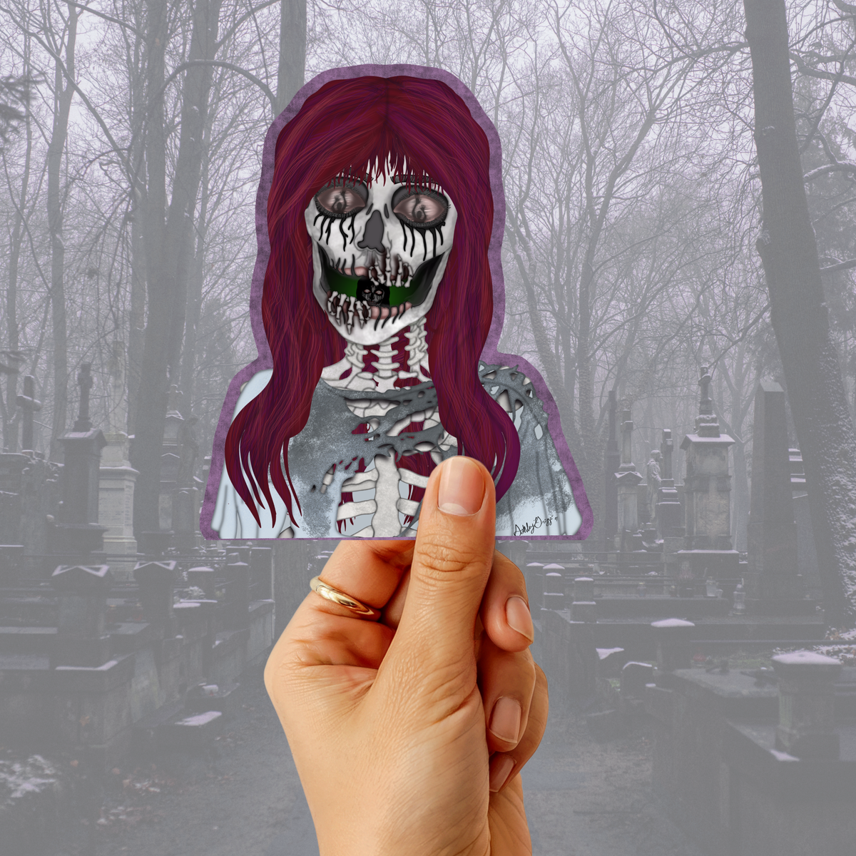 A hand holds a die-cut sticker featuring a skeleton character with long dark purple hair and a white shirt, set against a foggy cemetery background.