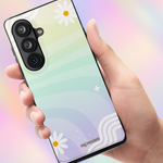 A hand holding a smartphone case with a pastel gradient design featuring white daisy flowers, wavy lines, and a black camera cutout, set against a soft rainbow-colored background.