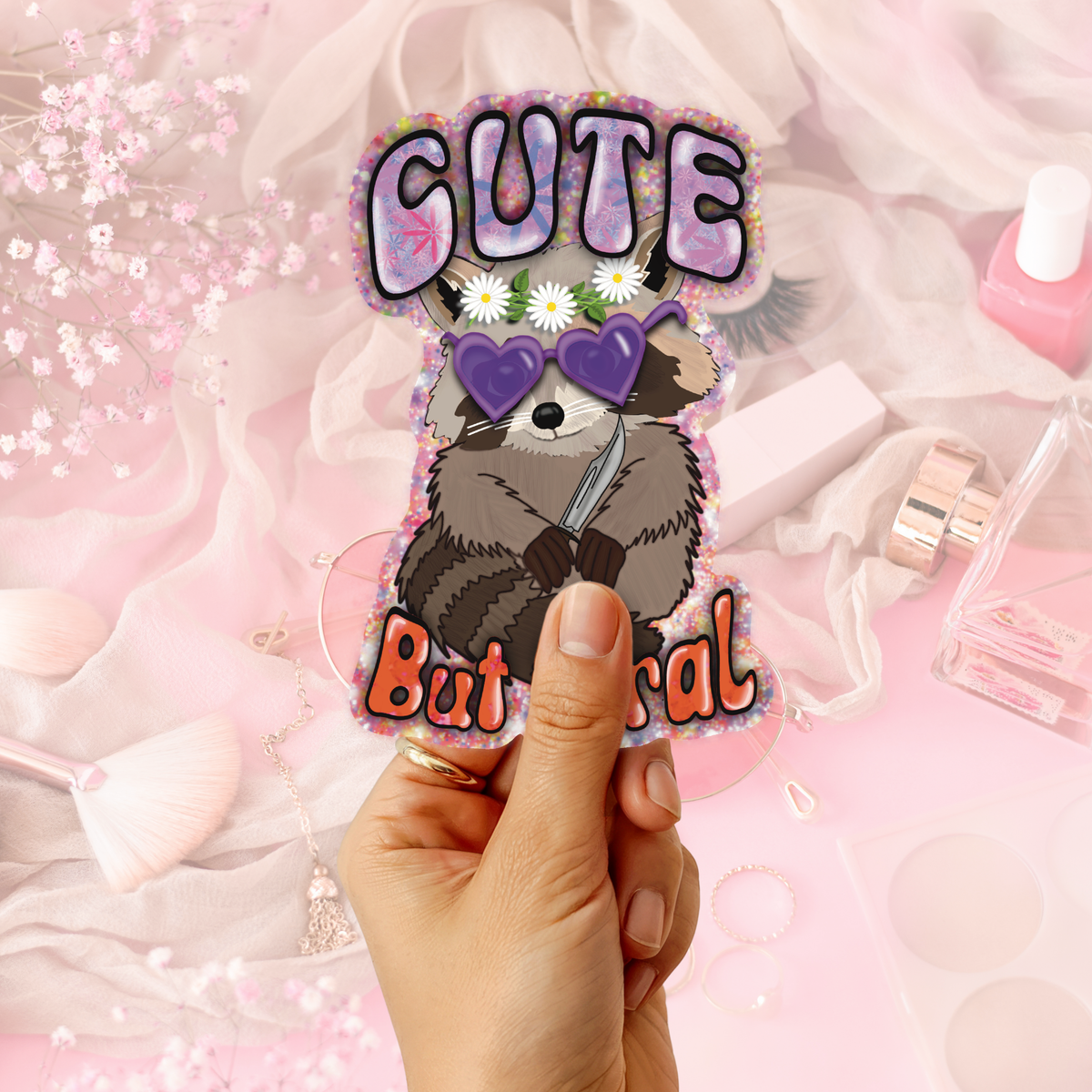 A hand holds a die-cut sticker featuring a cartoon raccoon wearing purple heart-shaped sunglasses, with the text 'CUTE But mental' in pink and orange lettering. The background is a styled flat-lay with pink cosmetics, sheer white fabric, and small flowers.