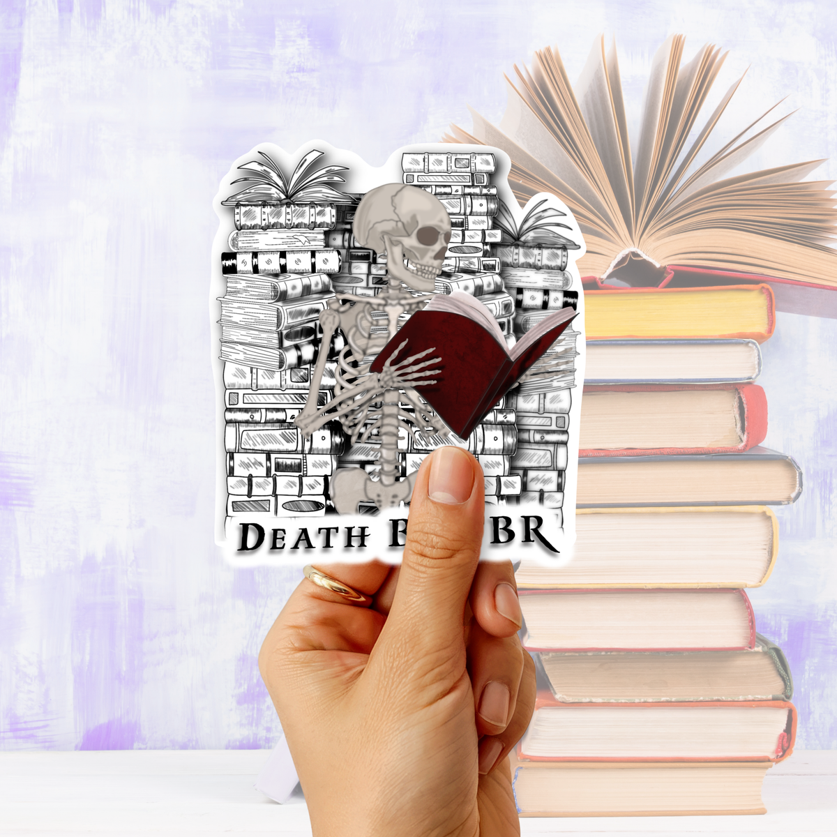 A hand holds a die-cut sticker featuring a black and white skeleton reading a dark red book in front of a bookshelf, with the text 'DEATH BY TBR' visible. The background shows a stack of books and an open book against a soft lavender-blue gradient backdrop.