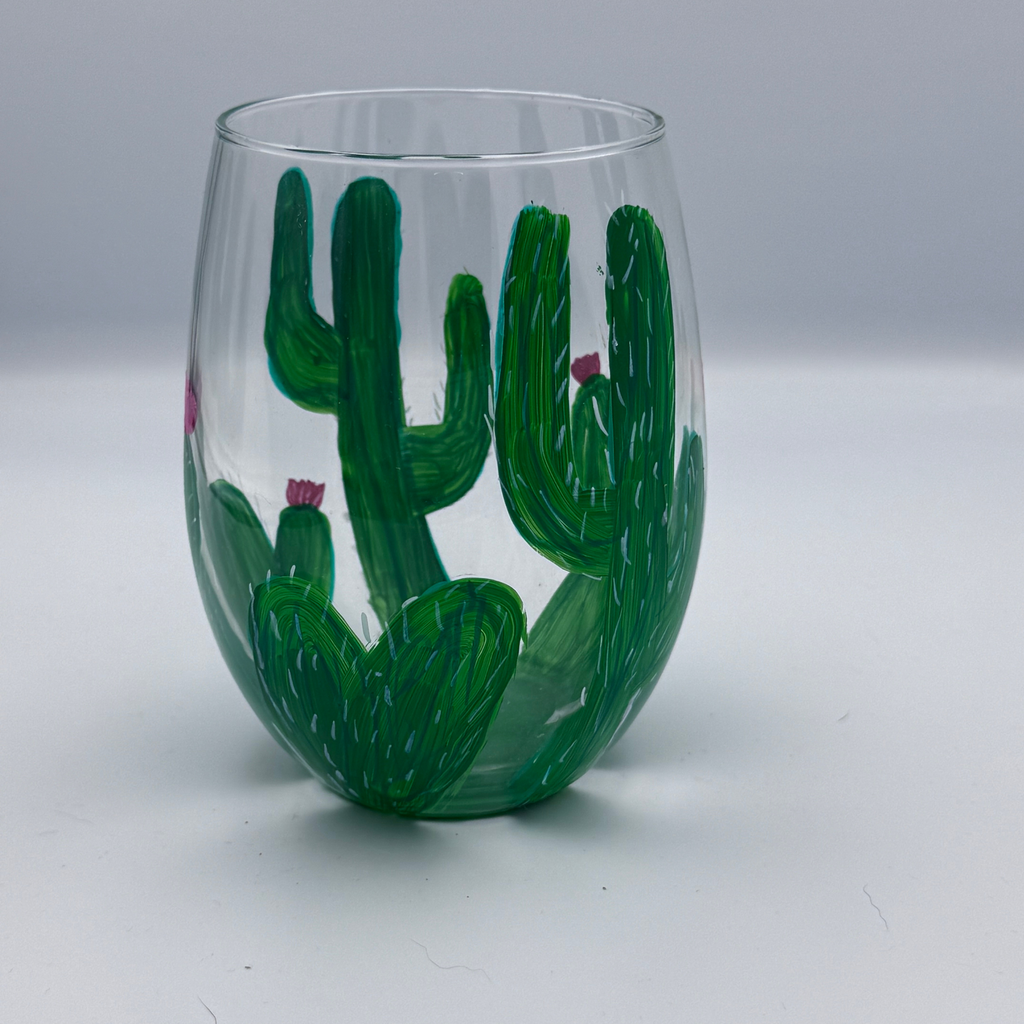 A clear stemless wine glass featuring a hand-painted green cactus design with small pink flowers, set against a neutral gray background.