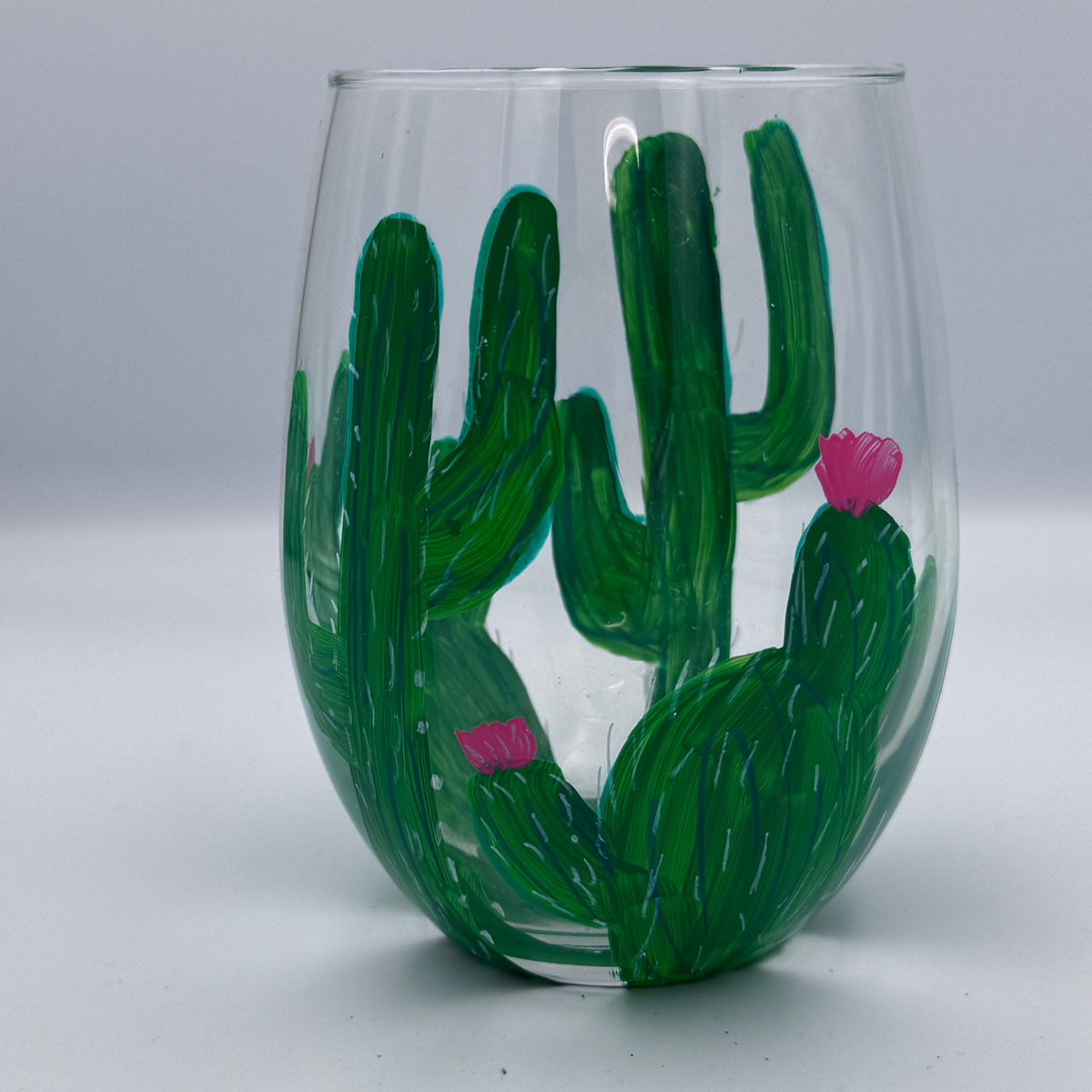 A clear stemless wine glass painted with green cacti and small pink flowers, shown against a light gray studio background.