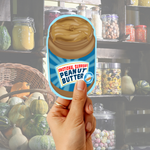 Hand holding an “Emotional Support Peanut Butter” sticker in front of a cozy pantry shelf background.