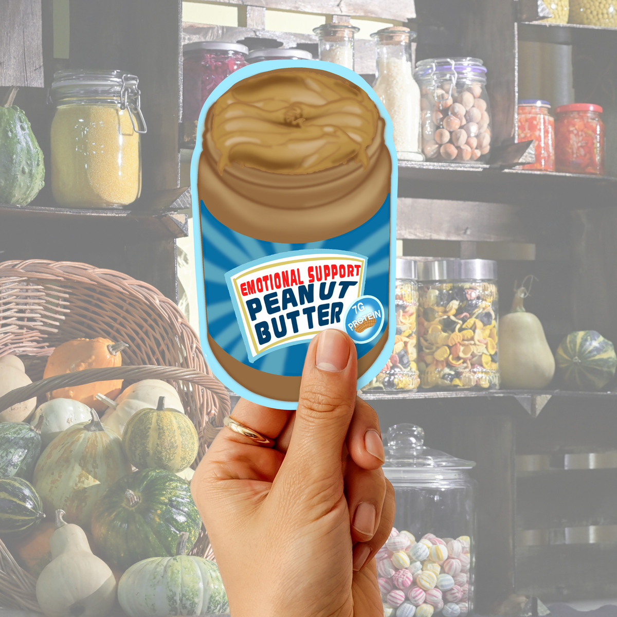 Hand holding an “Emotional Support Peanut Butter” sticker in front of a cozy pantry shelf background.