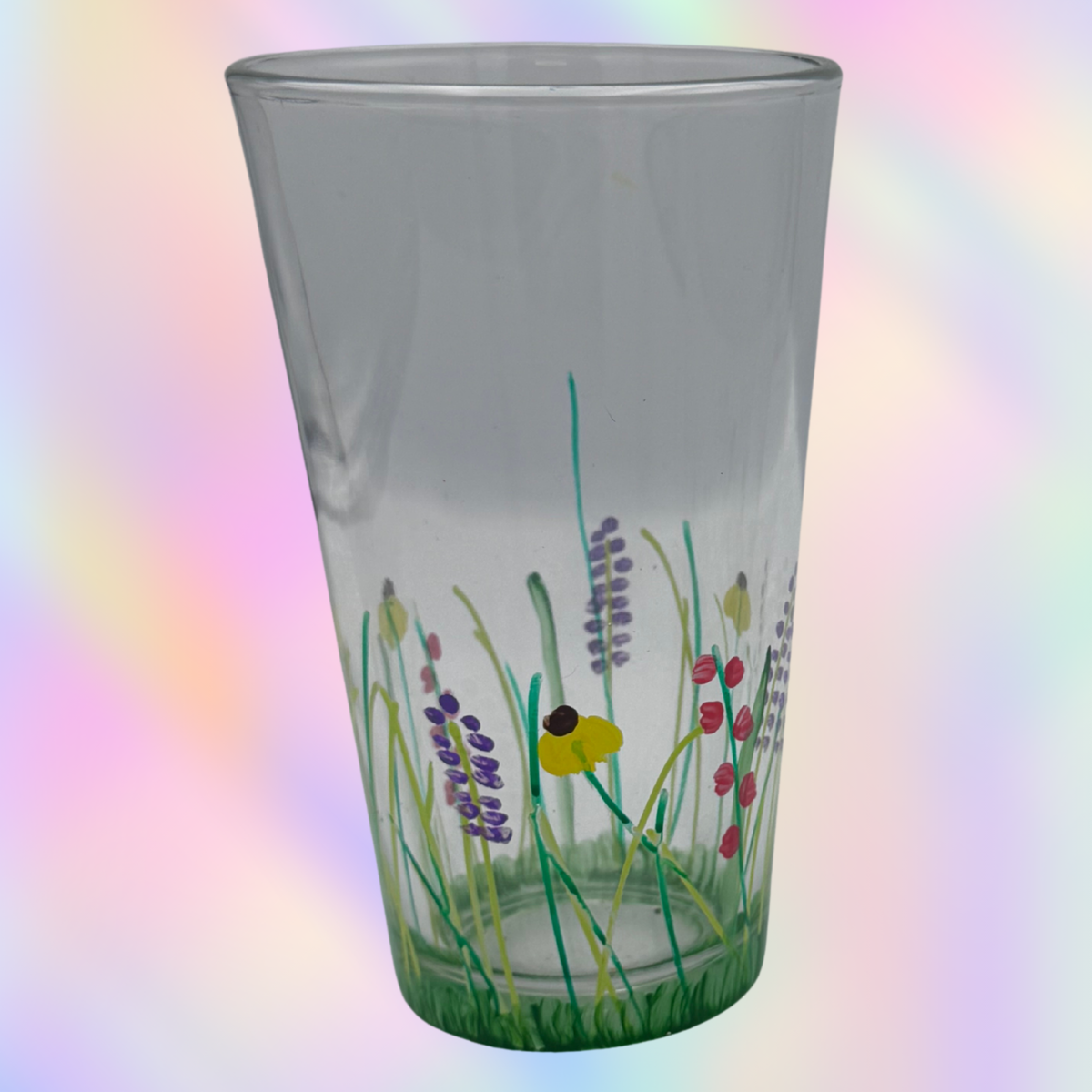 A clear glass pint glass decorated with a hand-painted wildflower and bee design near the base, set against a soft pastel rainbow gradient background.
