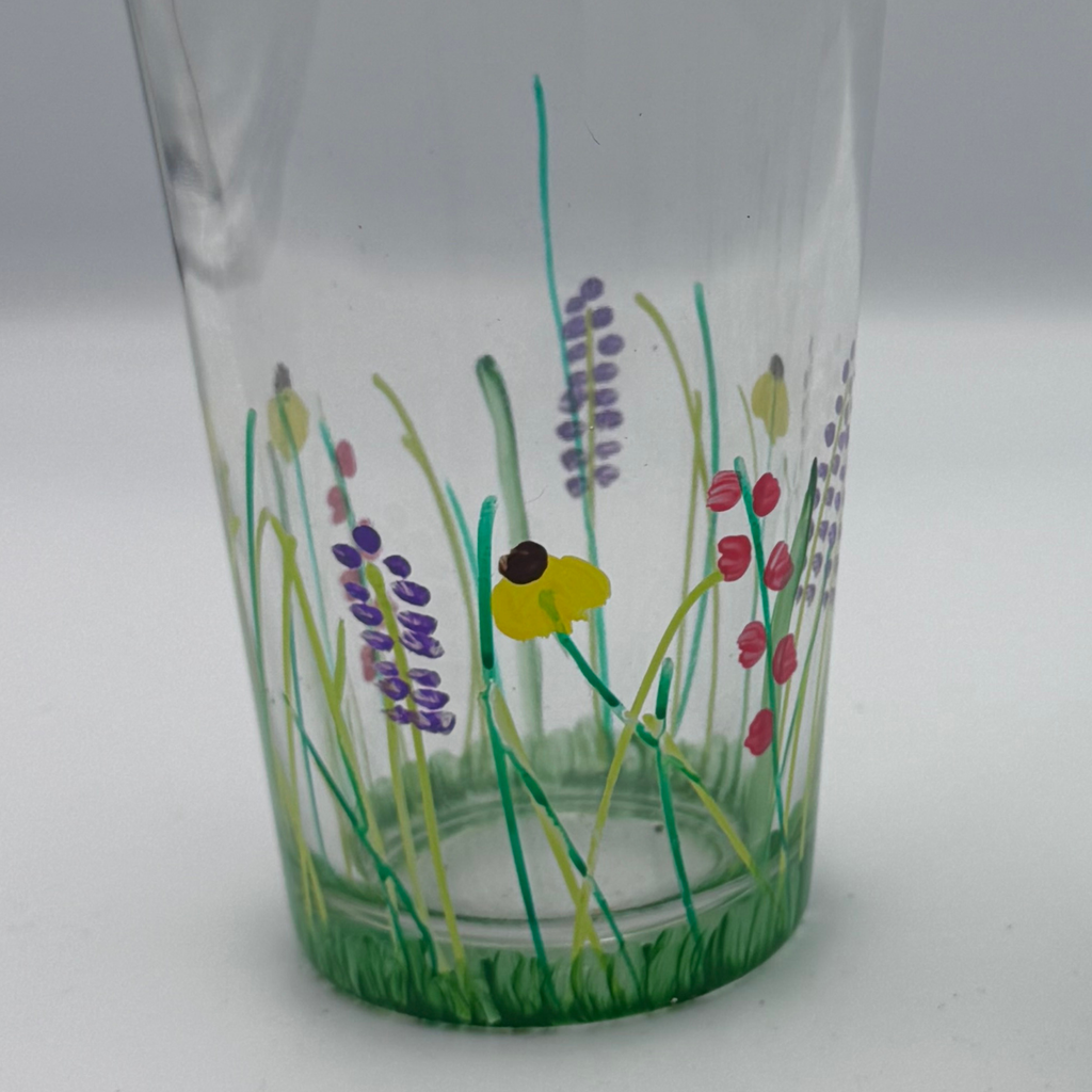 A clear glass drinking cup decorated with hand-painted grass and wildflowers, including green stems, purple lavender-like flowers, pink berries, and a yellow flower with a dark center, set against a neutral gray background.