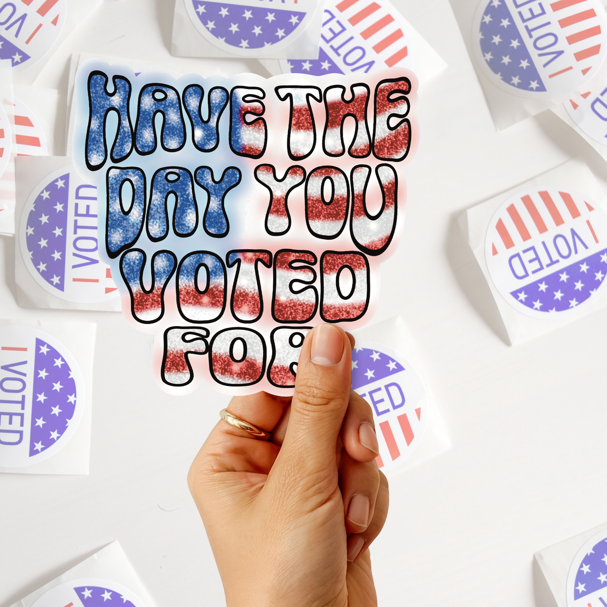 A hand holds a die-cut sticker that reads 'HAVE THE DAY YOU VOTED FOR?' in a red, white, and blue patriotic pattern. Surrounding the sticker are several circular stickers with 'I VOTED' in red, white, and blue.