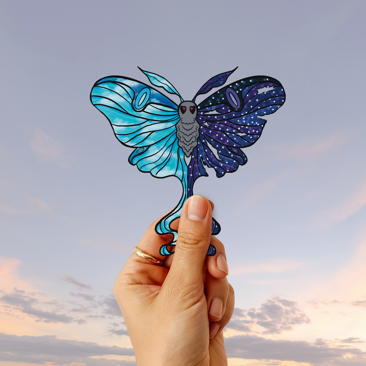 Hand holding a celestial luna moth inspired sticker against a soft sunset sky background.