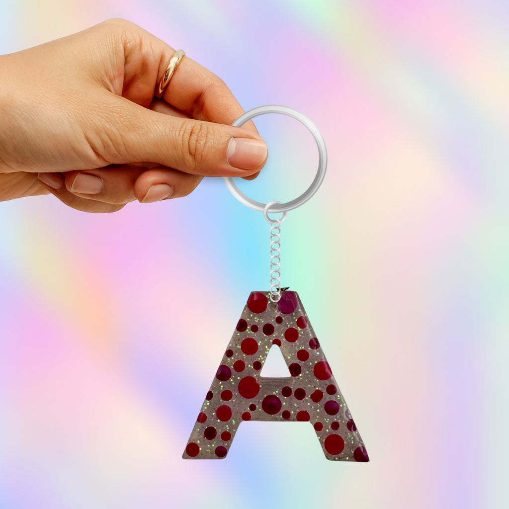 A hand holding a silver keychain with a glittery letter 'A' charm decorated with red and dark purple polka dots, set against a pastel rainbow gradient background.