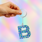 A hand holding a silver keyring with a dangling letter 'B' charm covered in blue and silver rhinestones, set against a pastel rainbow gradient background.