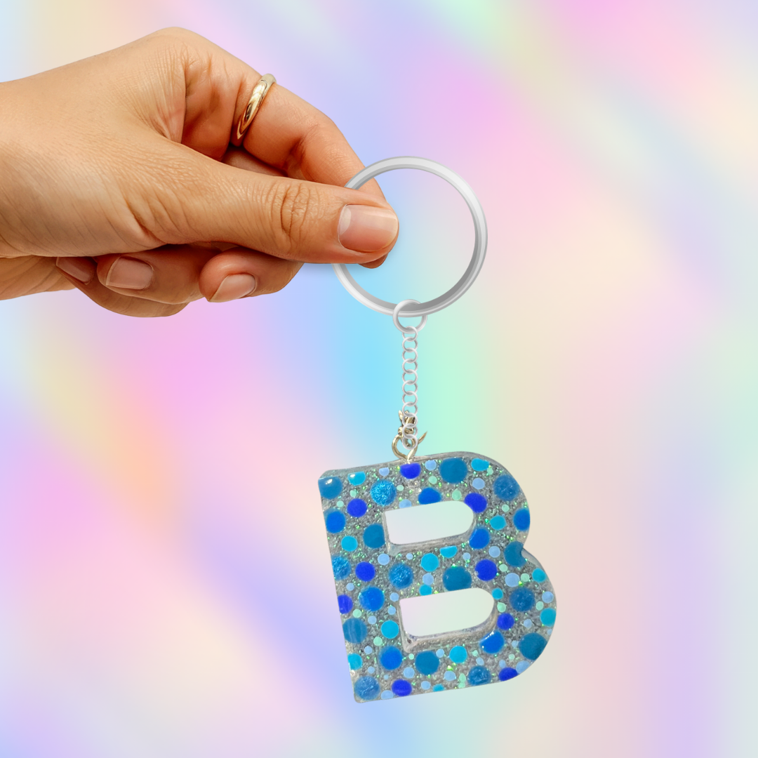 A hand holding a silver keyring with a dangling letter 'B' charm covered in blue and silver rhinestones, set against a pastel rainbow gradient background.