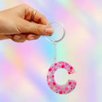 A hand holding a silver keyring with a dangling resin letter 'C' charm filled with pink, magenta, and silver glitter, set against a pastel rainbow iridescent background.