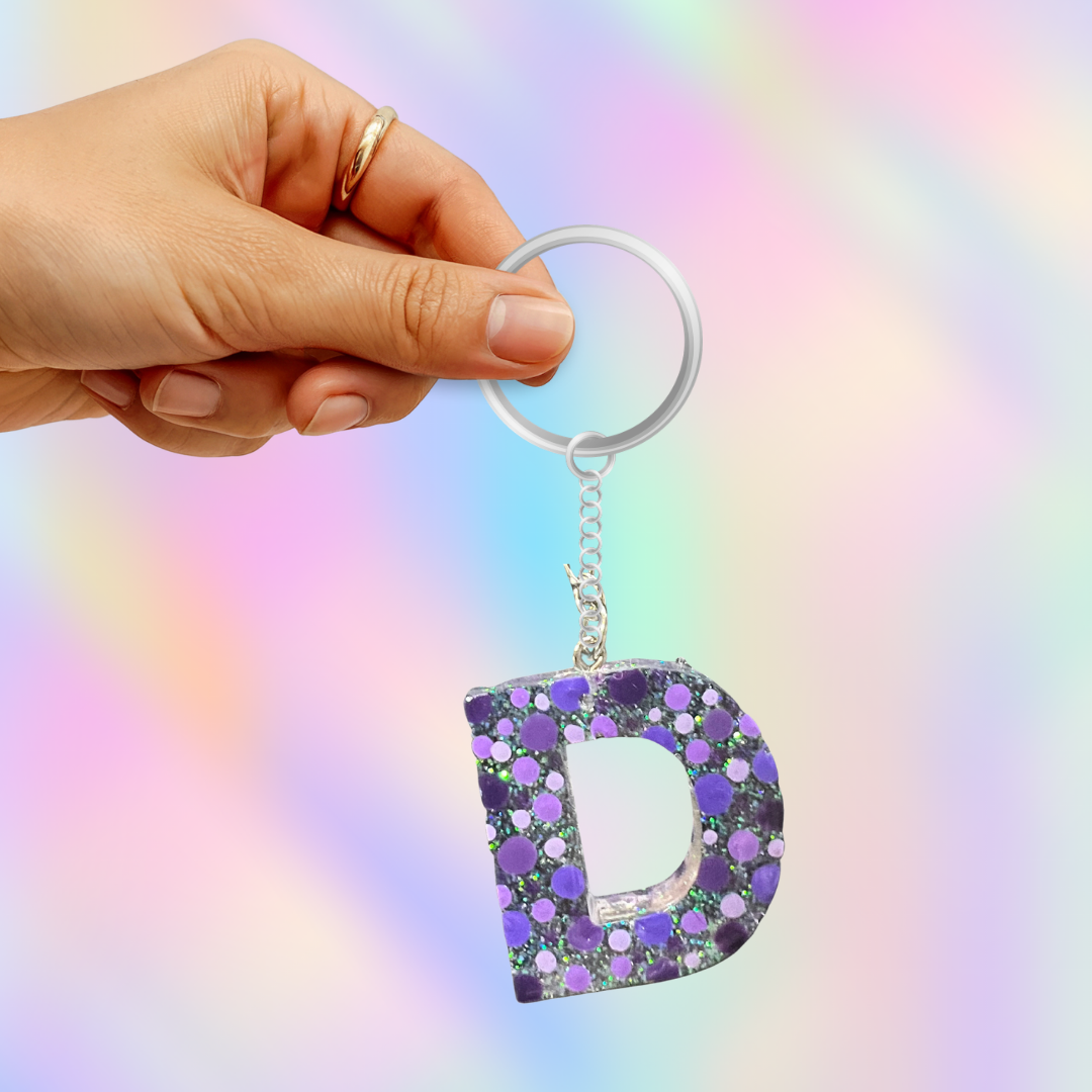 A hand holding a silver key ring with a glittery letter 'D' keychain in purple and silver, set against a pastel rainbow gradient background.