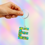 A hand holding a silver keychain with a glittery green letter 'E' charm covered in colorful green and yellow polka dots, set against a pastel rainbow gradient background.