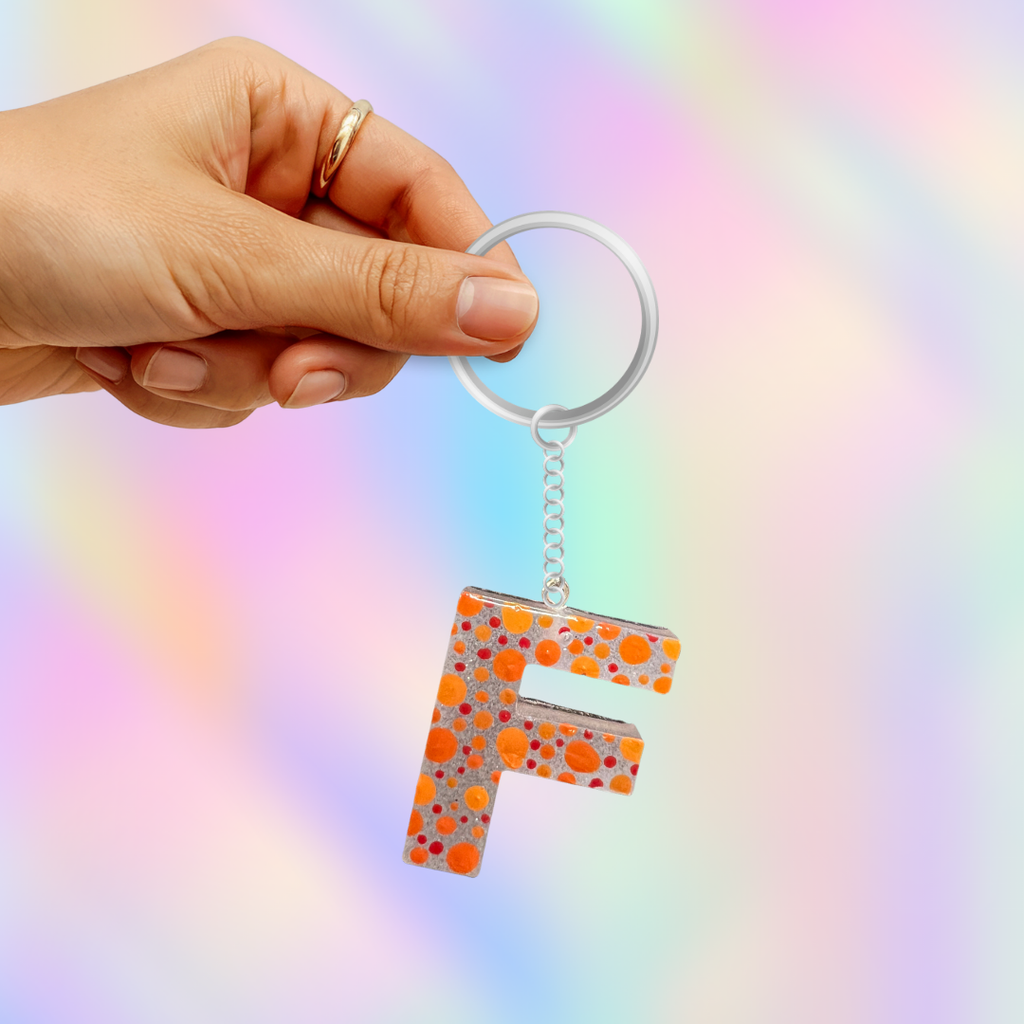 A hand holding a silver keyring with a letter 'F' acrylic keychain decorated with a colorful polka dot pattern of orange, pink, and blue on a pastel rainbow background.