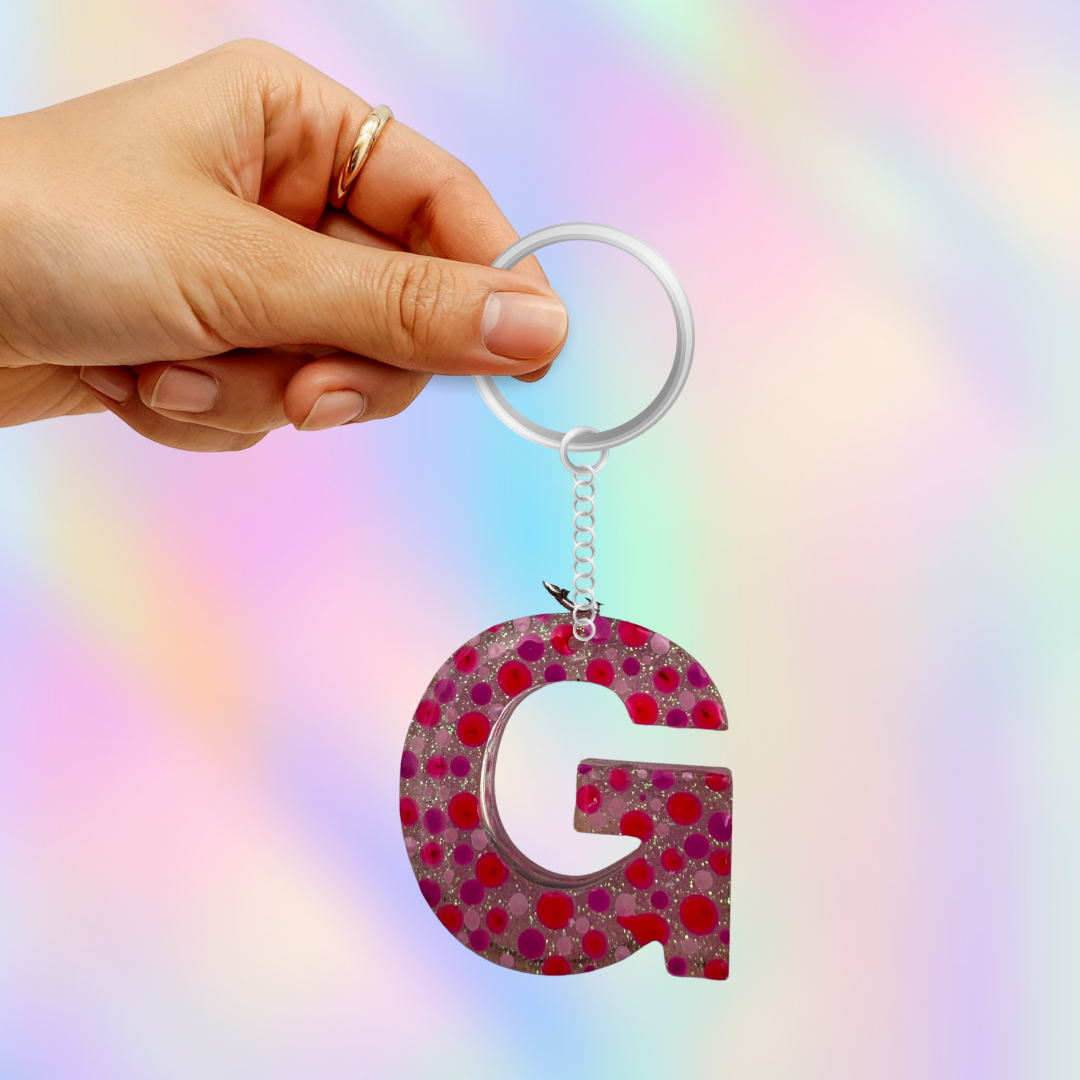 A hand holding a clear acrylic keyring with a glittery letter 'G' keychain featuring pink and red polka dots, set against a pastel rainbow gradient background.