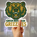 A hand holds a die-cut sticker featuring a green and gold grizzly bear head above the words 