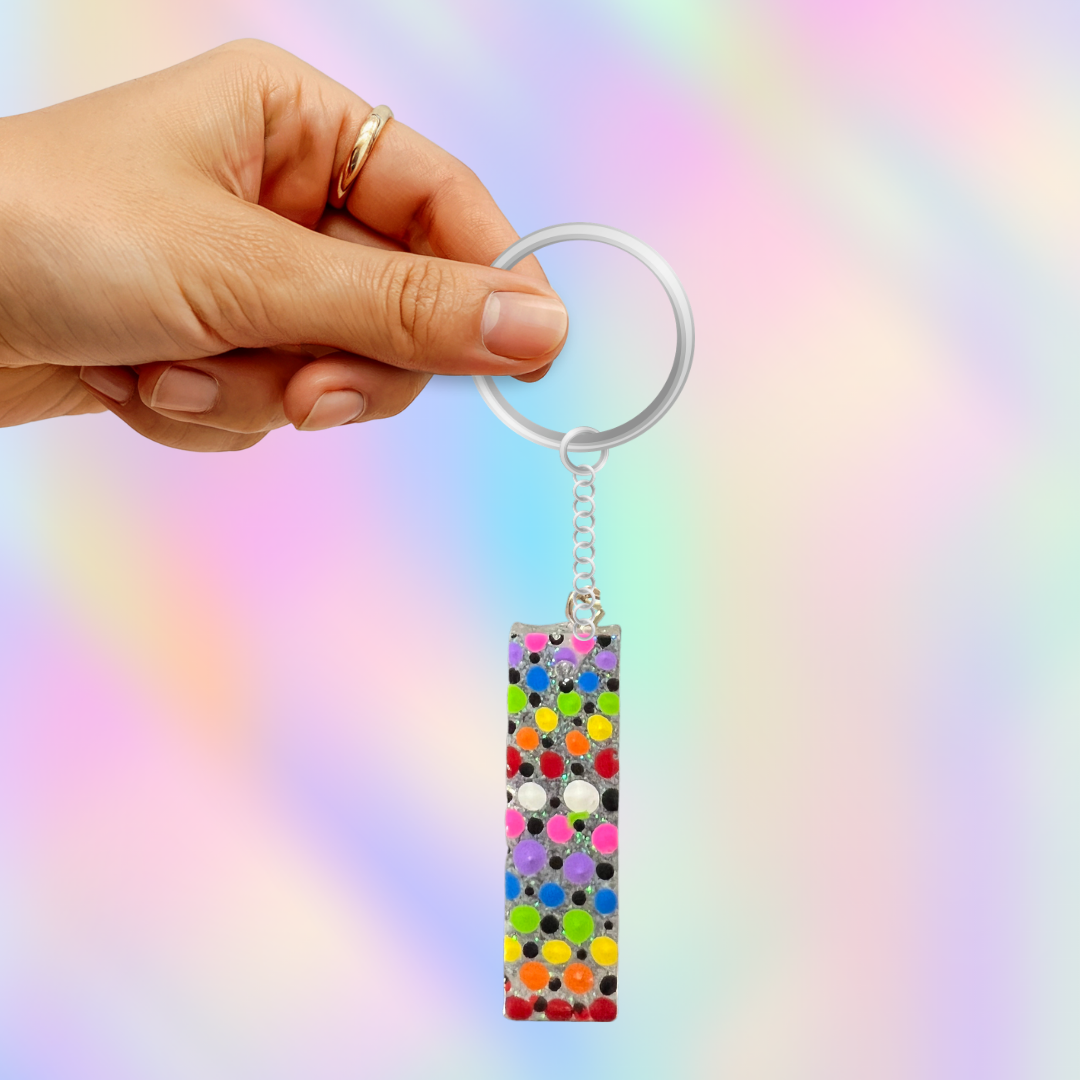 A hand holding a silver keychain with a rectangular clear acrylic charm covered in multicolored polka dots, set against a soft pastel rainbow gradient background.