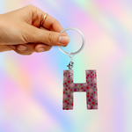 A hand holding a keychain with a translucent pink letter 'H' charm covered in colorful polka dots, attached to a white split ring, against a pastel rainbow gradient background.