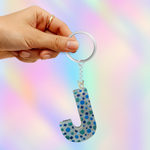 A hand holding a silver keyring with a glittery letter 'J' keychain featuring blue and teal polka dots on a light gray background with a pastel rainbow bokeh effect.