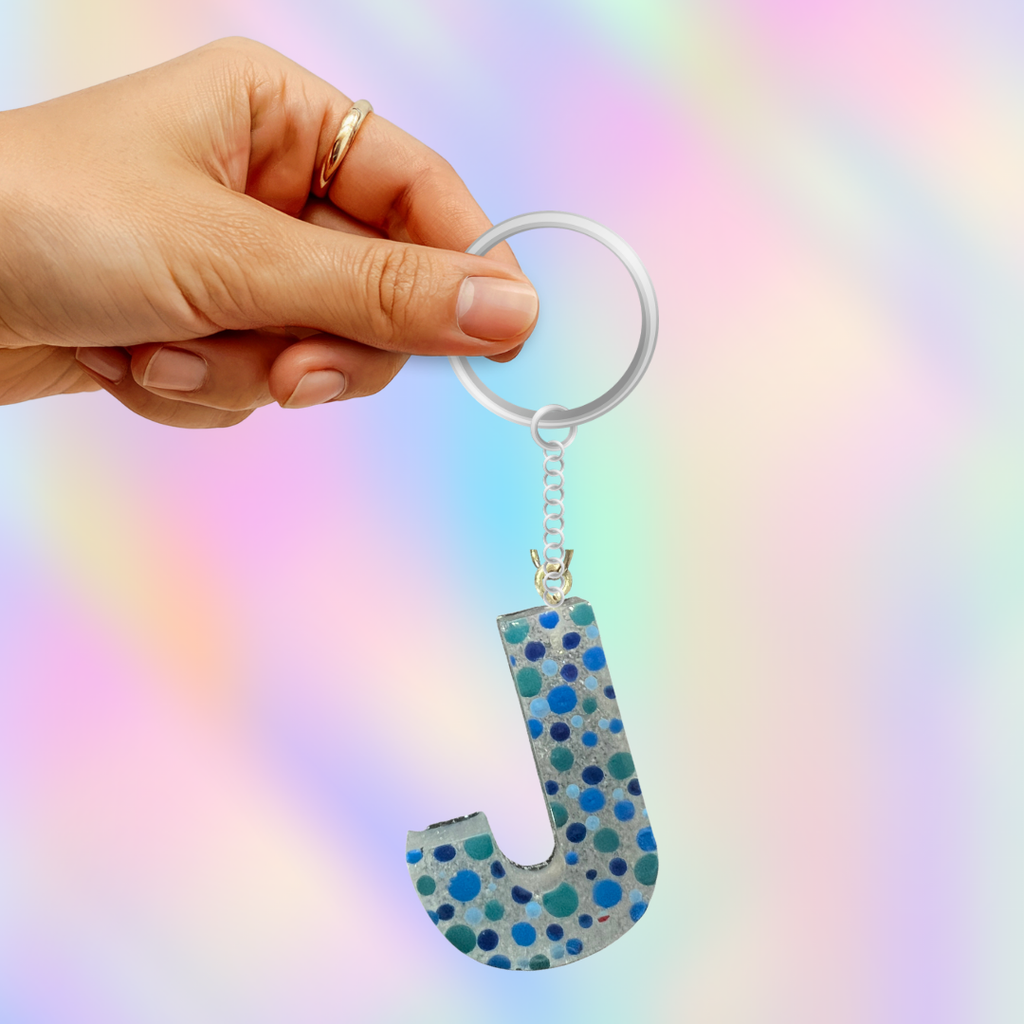 A hand holding a silver keyring with a glittery letter 'J' keychain featuring blue and teal polka dots on a light gray background with a pastel rainbow bokeh effect.
