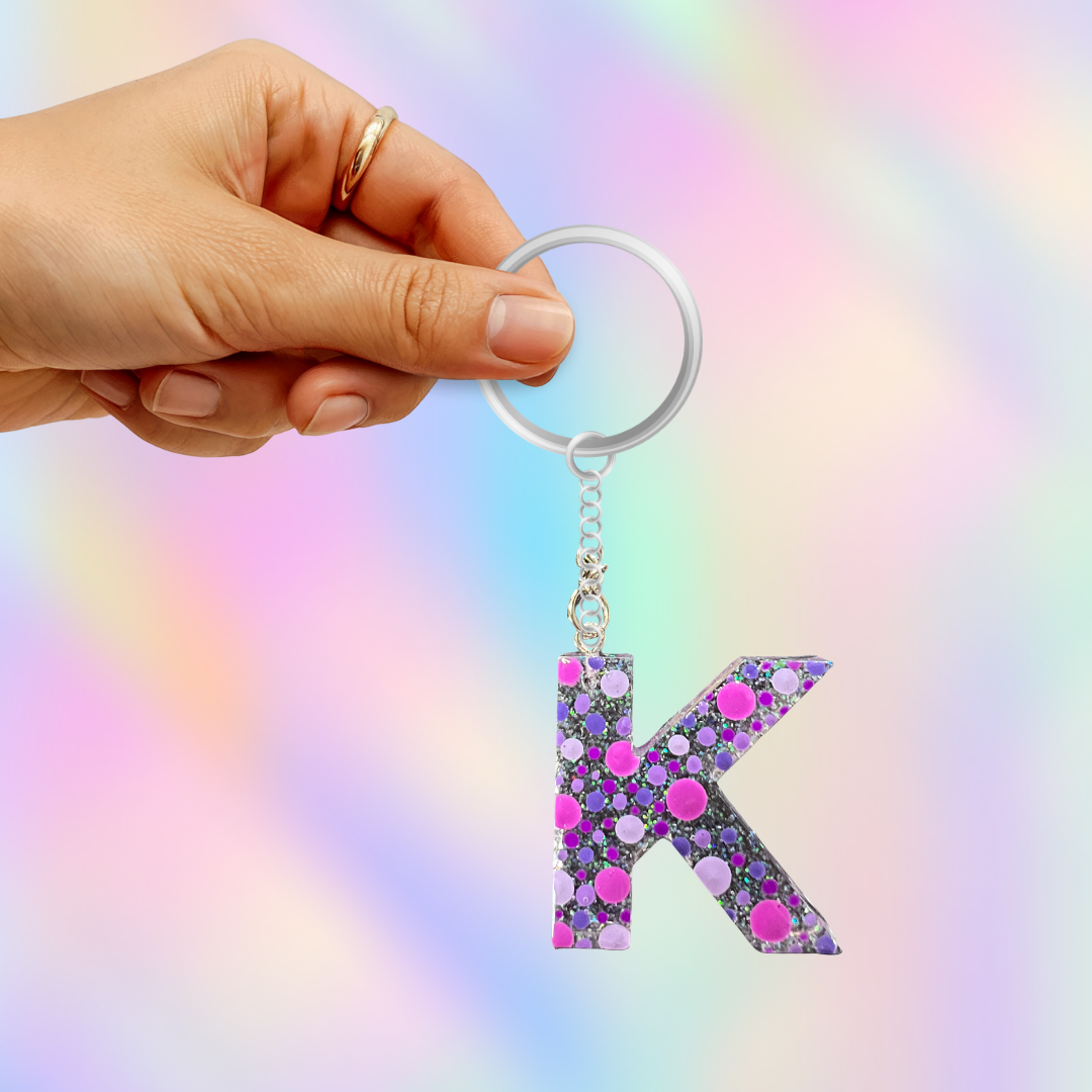 A hand holding a silver keyring with a dangling letter 'K' charm covered in pink and purple glitter, set against a pastel rainbow gradient background.