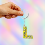 A hand holding a silver keyring with a dangling letter 'L' keychain filled with green, yellow, and white glitter, set against a pastel rainbow gradient background.