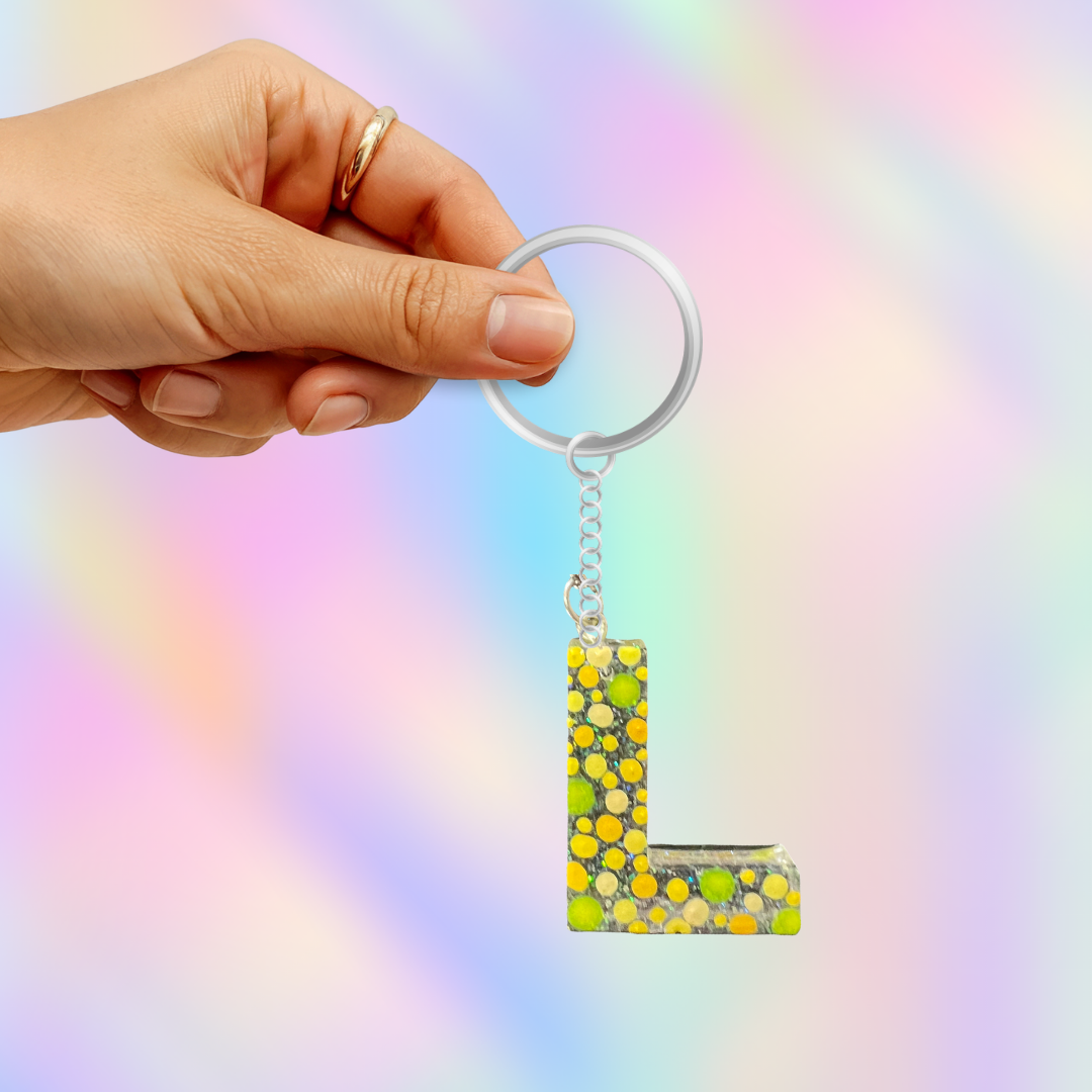 A hand holding a silver keyring with a dangling letter 'L' keychain filled with green, yellow, and white glitter, set against a pastel rainbow gradient background.