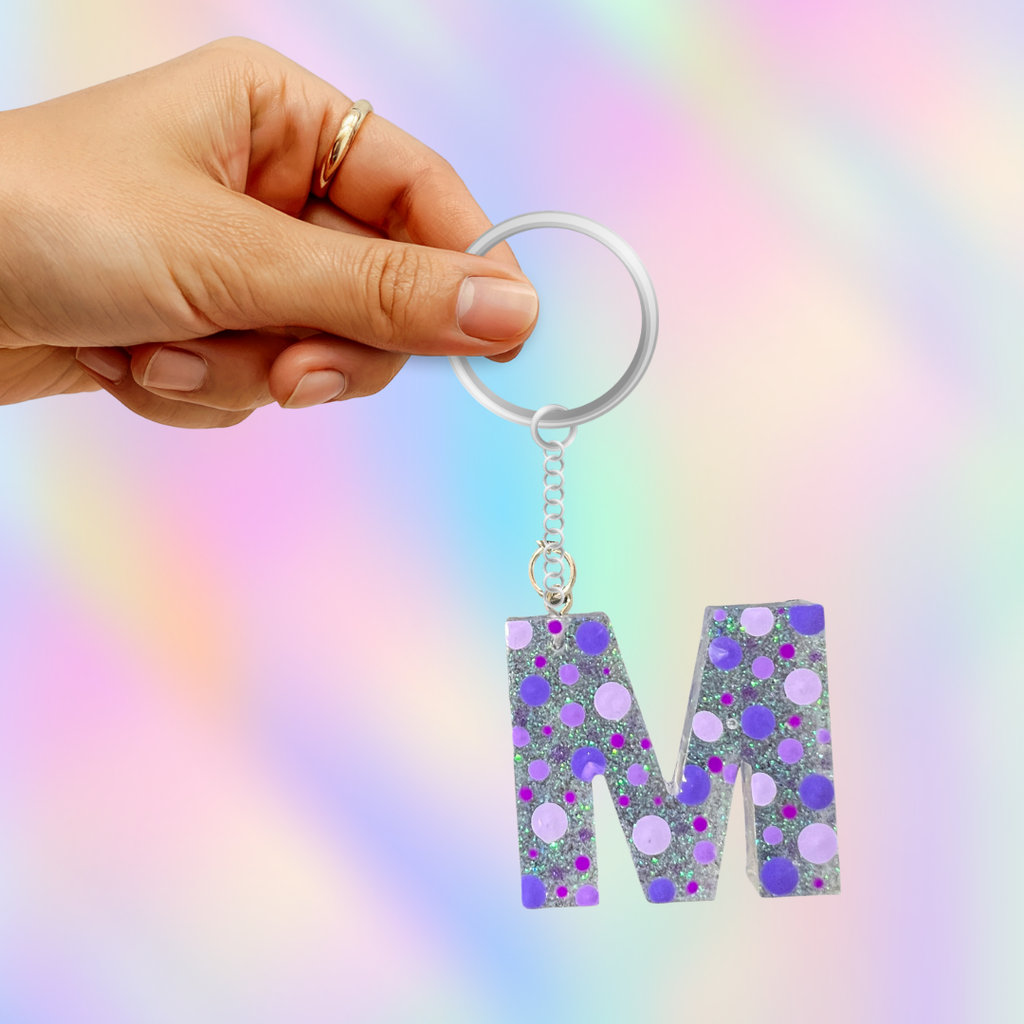 A hand holding a silver keyring with a dangling letter 'M' keychain. The letter 'M' is filled with silver glitter and decorated with purple and lavender polka dots, set against a pastel rainbow gradient background.