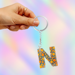 A hand holding a silver keyring with a dangling letter 'N' charm covered in orange and yellow floral details on a pastel rainbow background.