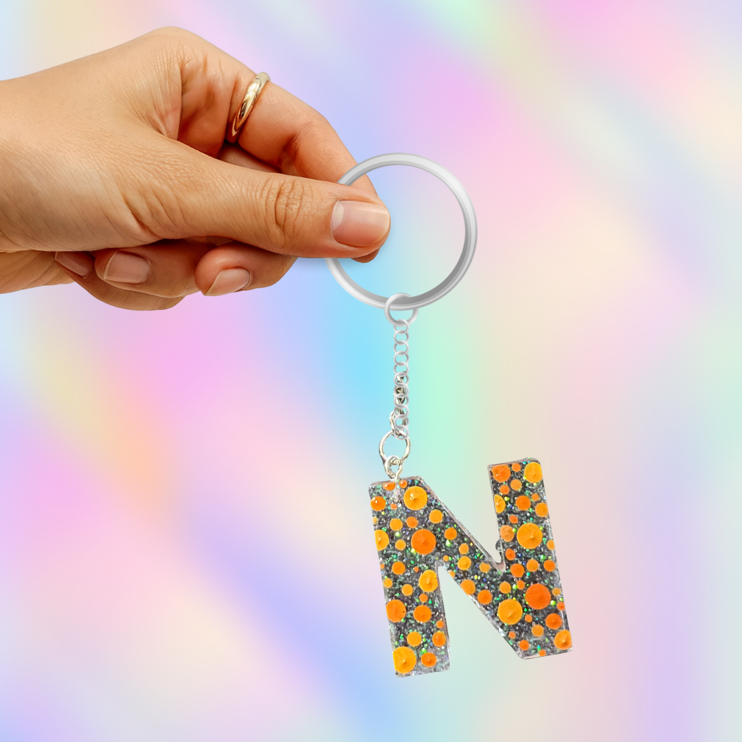 A hand holding a silver keyring with a dangling letter 'N' charm covered in orange and yellow floral details on a pastel rainbow background.