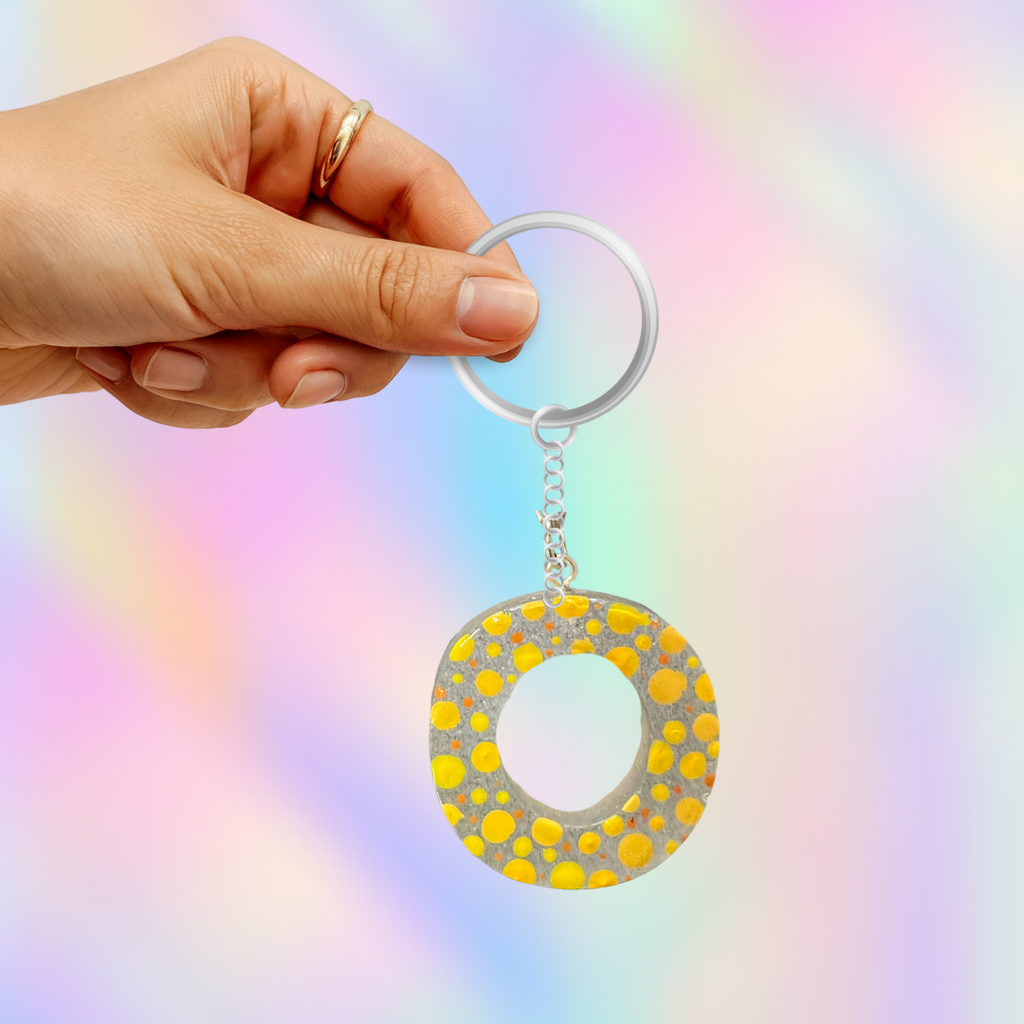 A hand holding a silver keychain with a circular acrylic charm decorated with yellow polka dots on a glittery silver background, set against a pastel rainbow gradient backdrop.