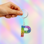 A hand holding a silver keyring with a colorful glittery letter 'P' charm, set against a pastel rainbow gradient background.