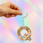 A hand holding a silver keyring with a dangling letter Q charm filled with orange and black glitter dots, set against a pastel rainbow gradient background.