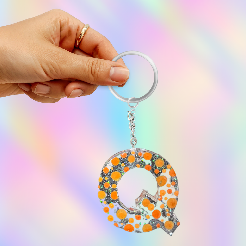 A hand holding a silver keyring with a dangling letter Q charm filled with orange and black glitter dots, set against a pastel rainbow gradient background.