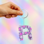 A hand holding a silver keyring with a glittery letter 'R' keychain in pink, purple, and silver colors, set against a pastel rainbow gradient background.