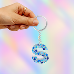A hand holding a clear acrylic key ring with a dangling letter 'S' charm filled with blue and light blue glitter, set against a pastel rainbow gradient background.