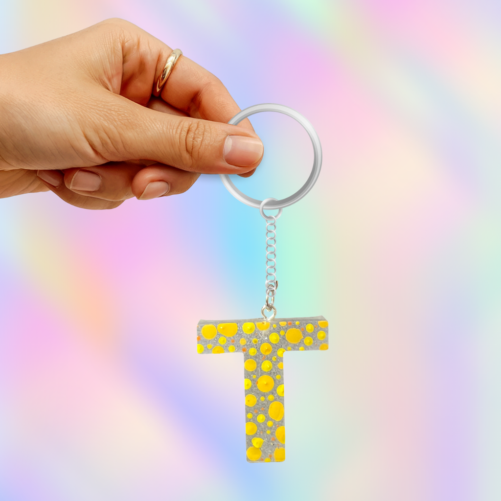 A hand holding a silver keyring with a yellow and silver glittery letter 'T' keychain against a pastel rainbow background.
