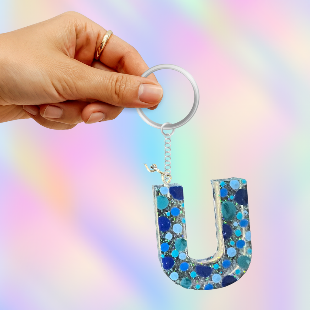 A hand holding a silver key ring with a glittery letter U keychain in shades of blue, teal, and silver against a pastel rainbow background.