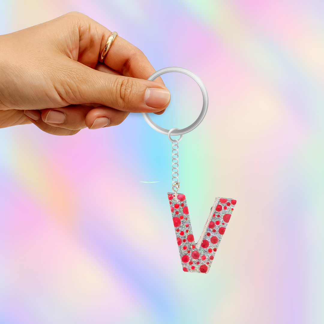 A hand holding a silver keyring with a dangling letter 'V' charm covered in red hearts and silver rhinestones, set against a pastel iridescent background.