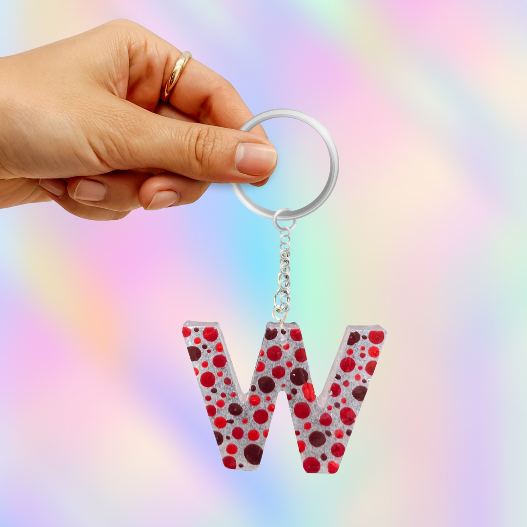 A hand holding a silver keyring with a dangling letter 'W' charm covered in red and dark red polka dots on a light gray background, set against a soft pastel rainbow gradient backdrop.