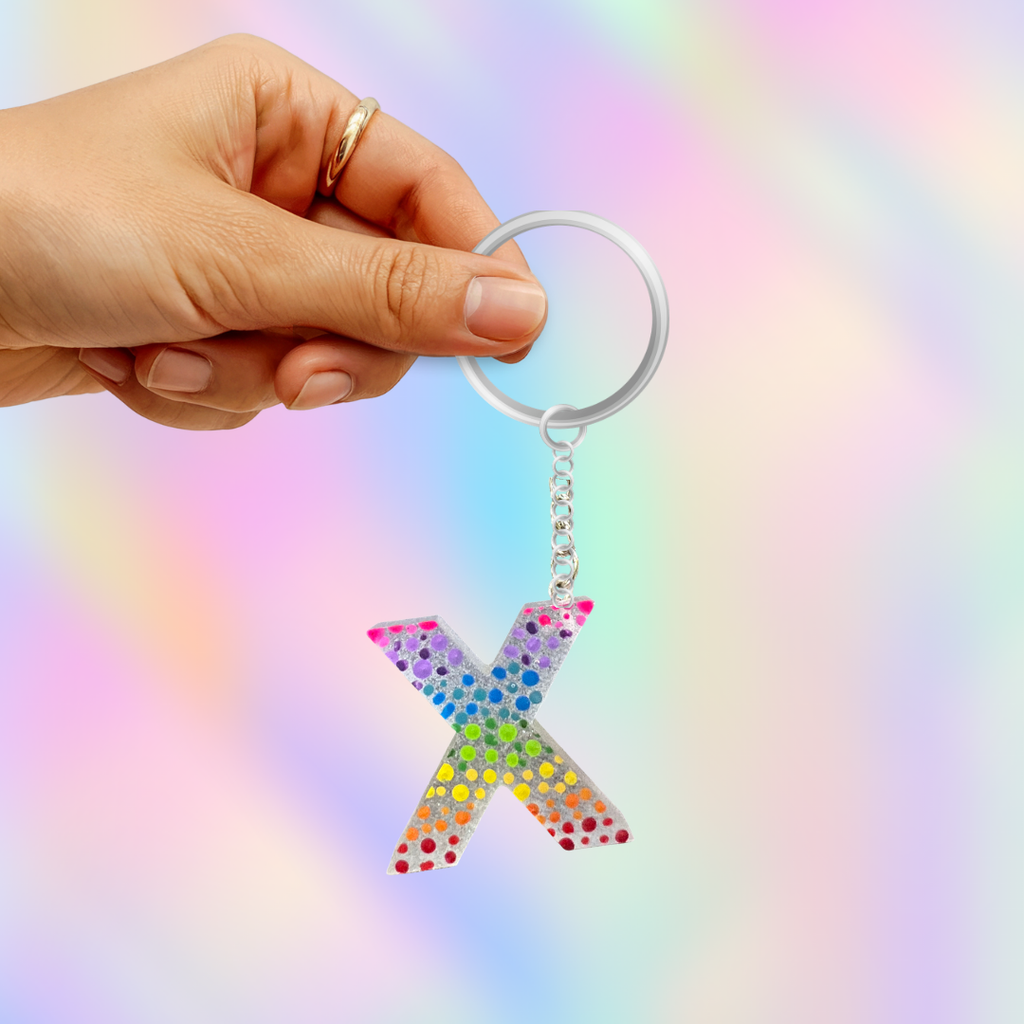 A hand holding a silver key ring with a glittery rainbow 'X' keychain against a pastel iridescent background.