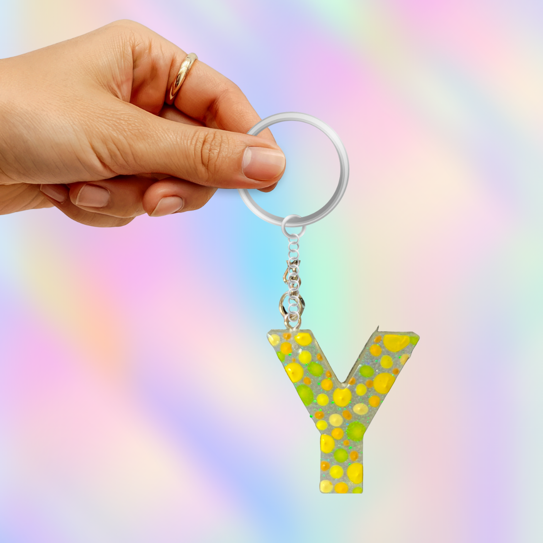 A hand holds a silver keyring with a dangling letter Y keychain. The letter Y is filled with a yellow and green polka dot pattern and hangs from a silver chain.