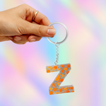 A hand holding a silver keyring with a dangling letter 'Z' keychain. The letter 'Z' is decorated with a pattern of orange glitter and silver rhinestones, set against a soft, iridescent pastel background.